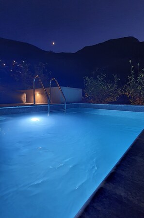 Une piscine éclairée la nuit, avec des échelles en métal, entourée de végétation et un bâtiment visible au loin.