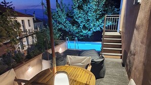 Een terras buiten met een houten tafel en stoelen, een hangende parasol en een uitzicht op een zwembad en gebouwen op de achtergrond.
