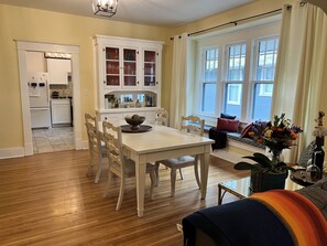 Un comedor con una mesa y sillas blancas, una cocina con muebles blancos y un gran ventanal con cortinas.