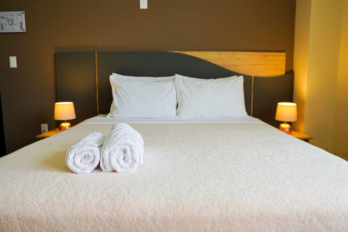 A neatly made bed with white linens and two rolled towels on the bed. There are two bedside lamps on either side of the bed.