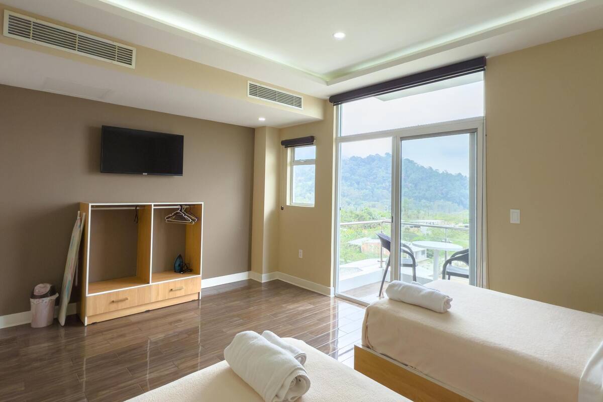 A hotel room with two beds, a TV, a large window, and a balcony with a view of the mountains.