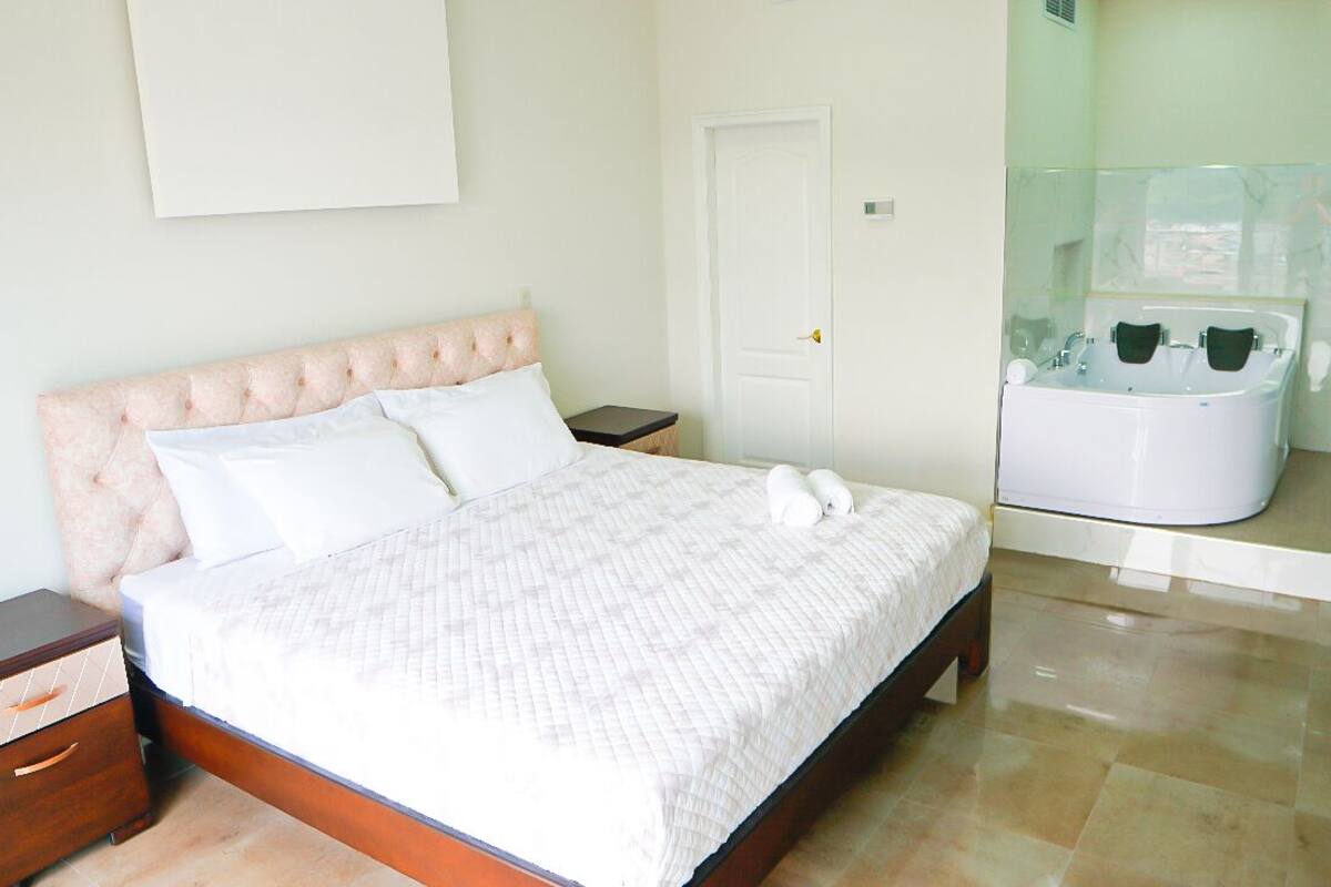 A bedroom with a large bed, a nightstand, a wooden dresser, and a glass-enclosed bathroom with a bathtub.