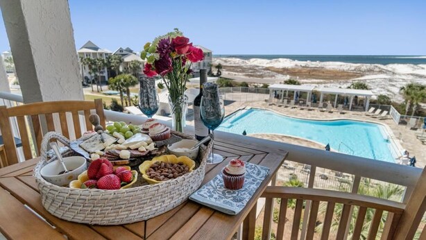 Un tavolo con un cesto di frutta, un bicchiere di vino e un dessert su un balcone con vista su una piscina e una spiaggia.