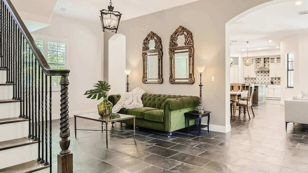 Un amplio salón con un sofá verde, dos espejos decorados, una escalera y una cocina al fondo.