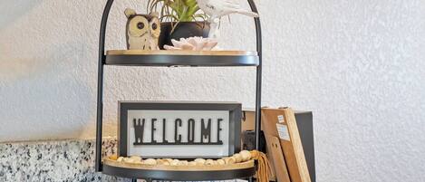 A tiered display stand with a welcome sign, a bird figurine, a plant, and a small owl figurine.