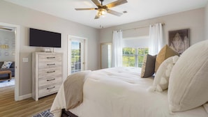 Une chambre à coucher avec un lit, un ventilateur de plafond, une télévision, une commode et une fenêtre avec des rideaux.