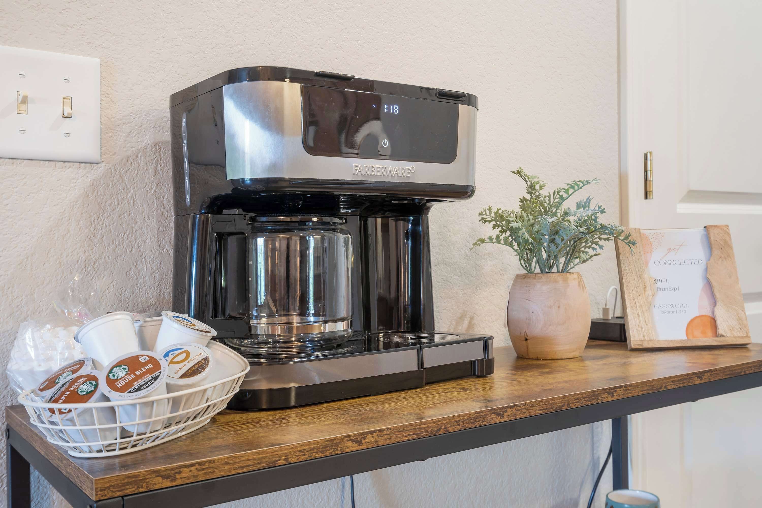 A Farberware coffee maker on a wooden counter with a basket of coffee cups and a potted plant.