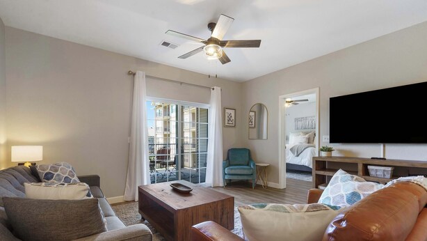 Un salon comprenant un canapé, un fauteuil, une table basse et un téléviseur. On y trouve également un ventilateur de plafond, une fenêtre avec des rideaux et une porte donnant sur une autre pièce.