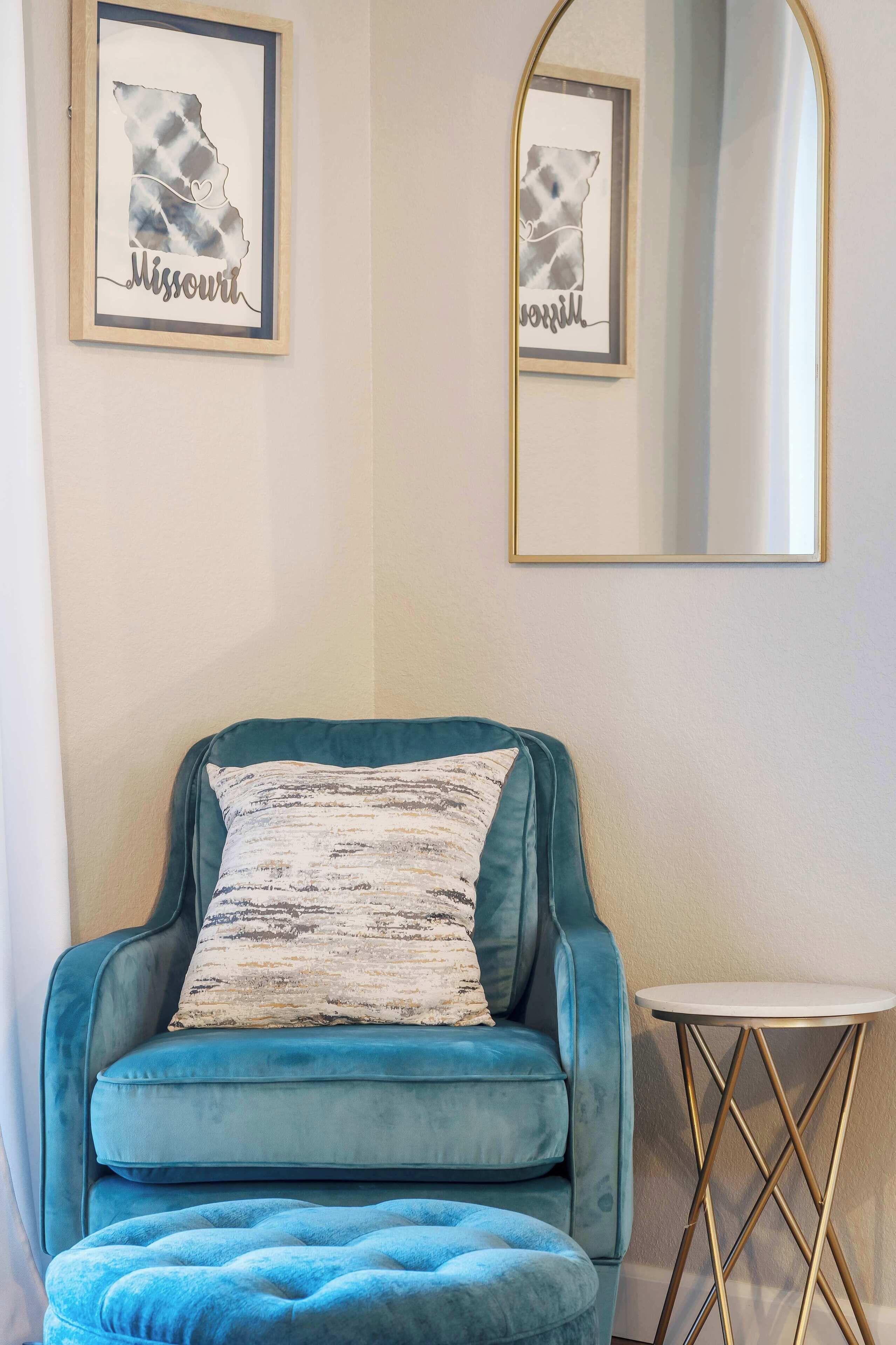 A teal armchair with a patterned pillow, a small round table, and two framed artworks on the wall.