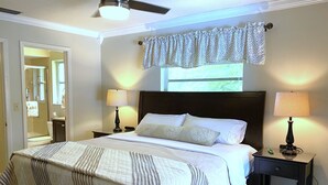 Una cama bien hecha con cubrecama a rayas, dos lámparas de noche, una ventana con cortina decorativa y un ventilador de techo.
