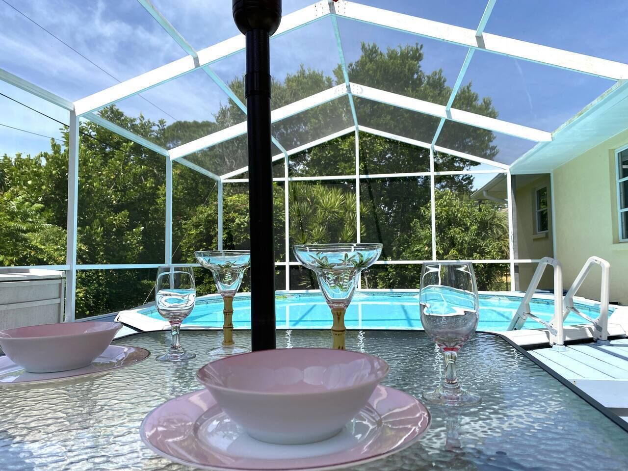 A glass-enclosed outdoor area with a pool, dining table with pink bowls and wine glasses, and a white railing.