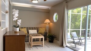 Amplia sala de estar con sofá, mesa de centro y ventilador de techo. Se ve una ventana grande con cortinas, una planta en maceta y vistas al exterior con vegetación.