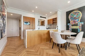 A modern kitchen with a marble countertop, wooden cabinets, and a dining area with a round table and chairs. - The Penthouse - Sebel (Auckland)