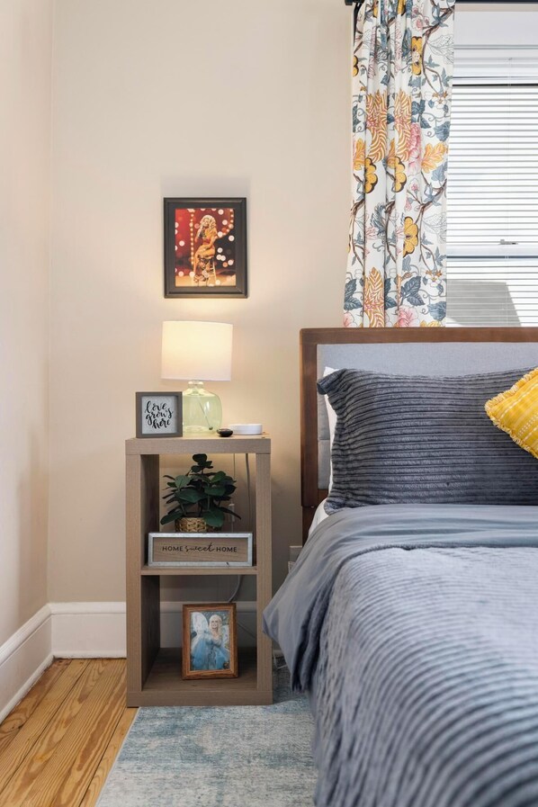 A bedroom with a bed, a nightstand with a lamp, a framed picture, a small shelf with a plant, and a window with curtains.