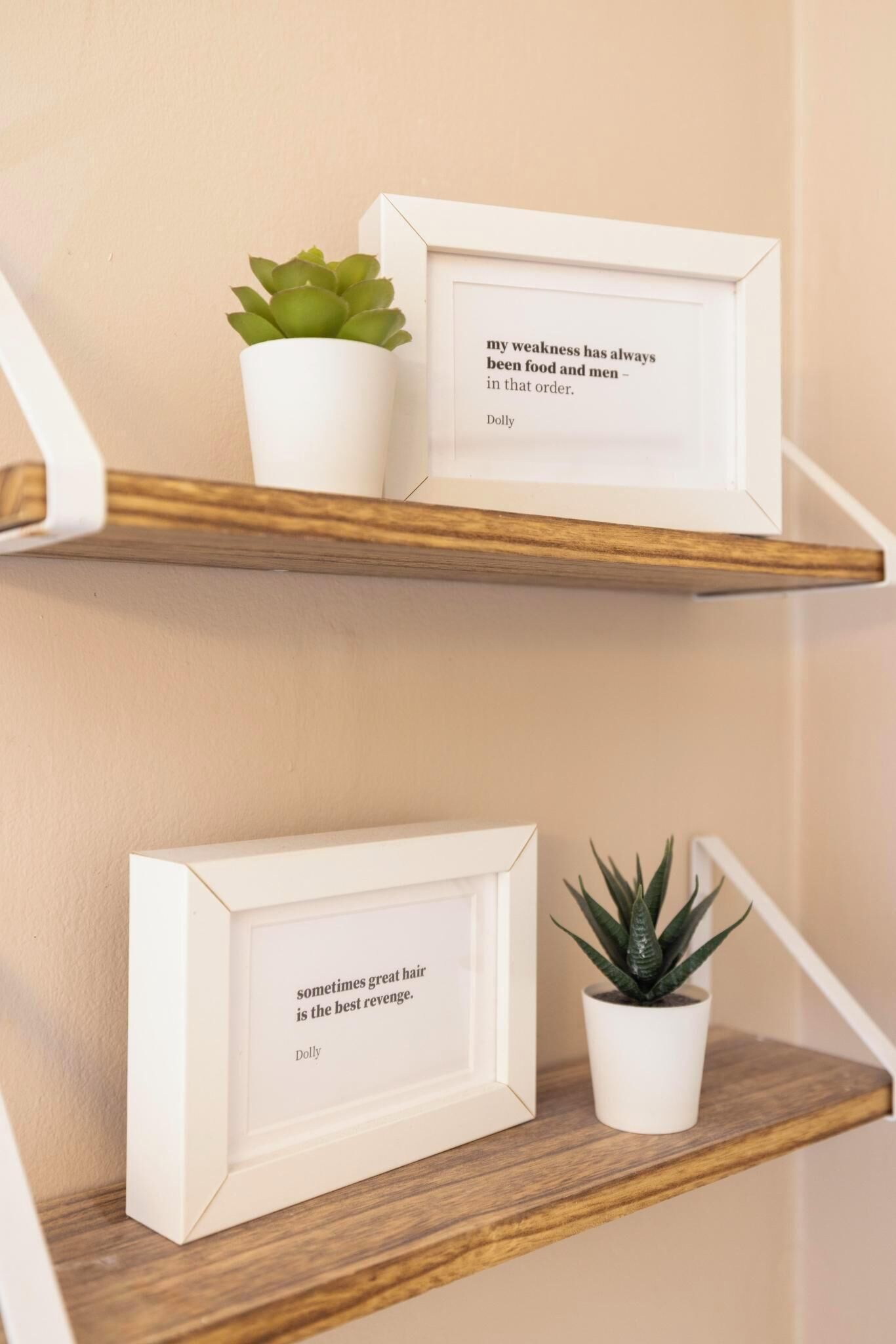 Twee witte fotolijstjes op houten planken tegen een muur, elk met een citaat van Dolly en een kleine potplant.