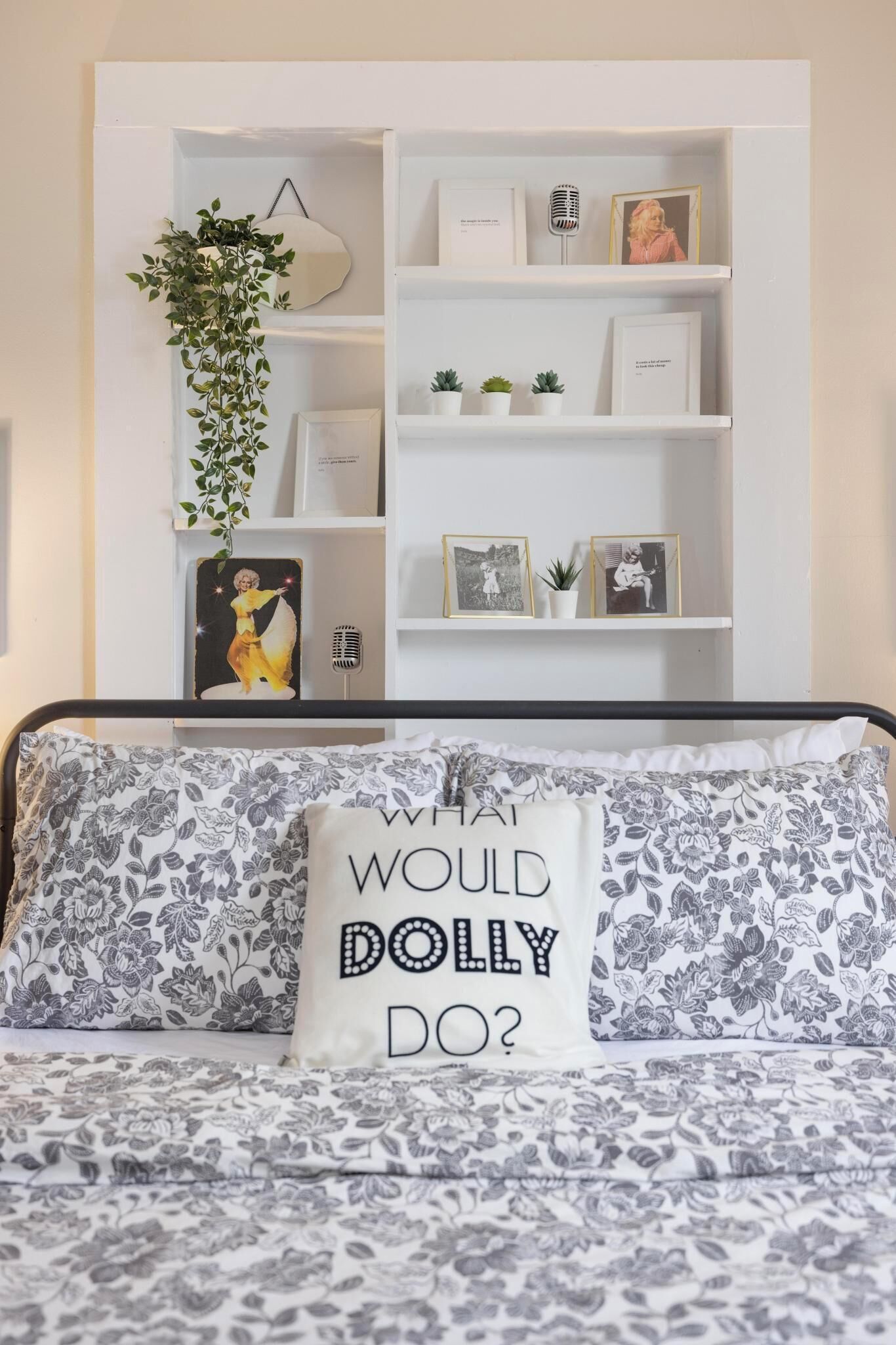 A bed with floral-patterned bedding and a pillow with text. Behind the bed, there is a white shelving unit with various items including books, framed photos, and small plants.