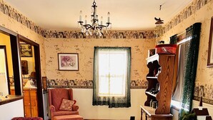 Une pièce avec un plancher en bois, un fauteuil rouge, une bibliothèque en bois, un lustre et une fenêtre ornée de papier peint à motifs.