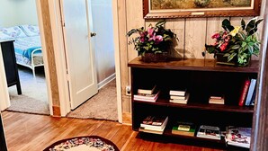 Une pièce avec un plancher en bois, un tapis à motifs floraux, une bibliothèque remplie de livres et une plante en pot, un tableau encadré et une porte portant l’inscription « ANTIQUITÉS ».