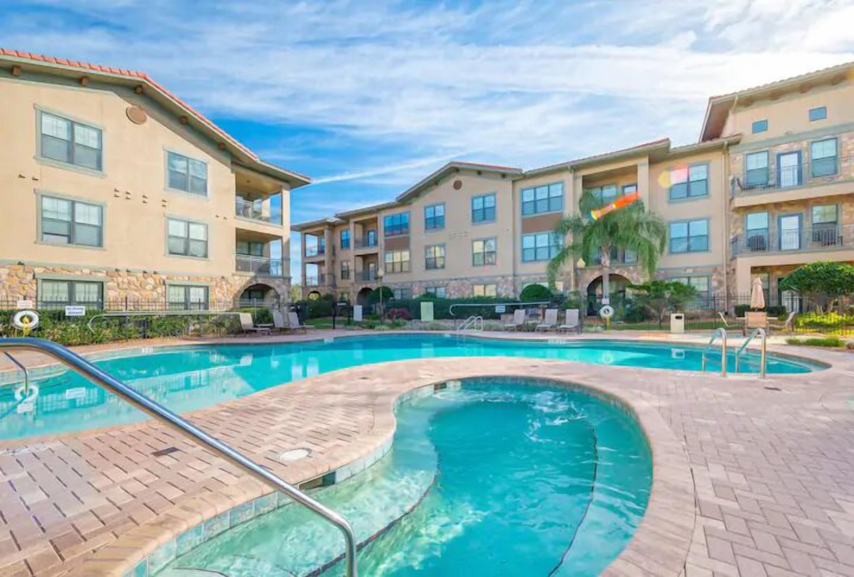 A swimming pool area with a brick deck, surrounded by multi-story buildings with balconies and windows.