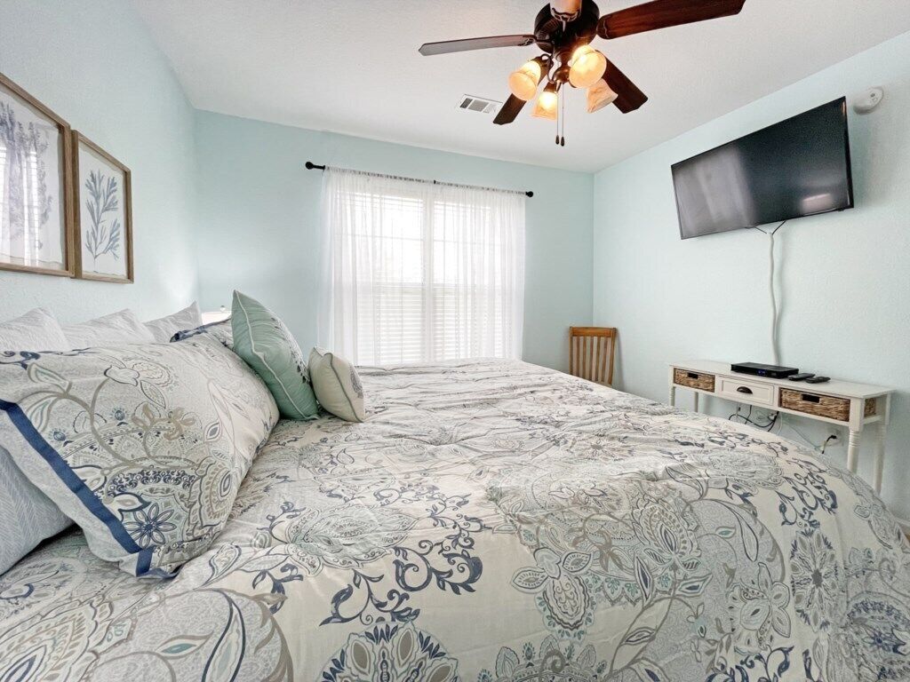 Un dormitorio con cama, ventilador de techo, televisor montado en la pared y ventana con cortinas.