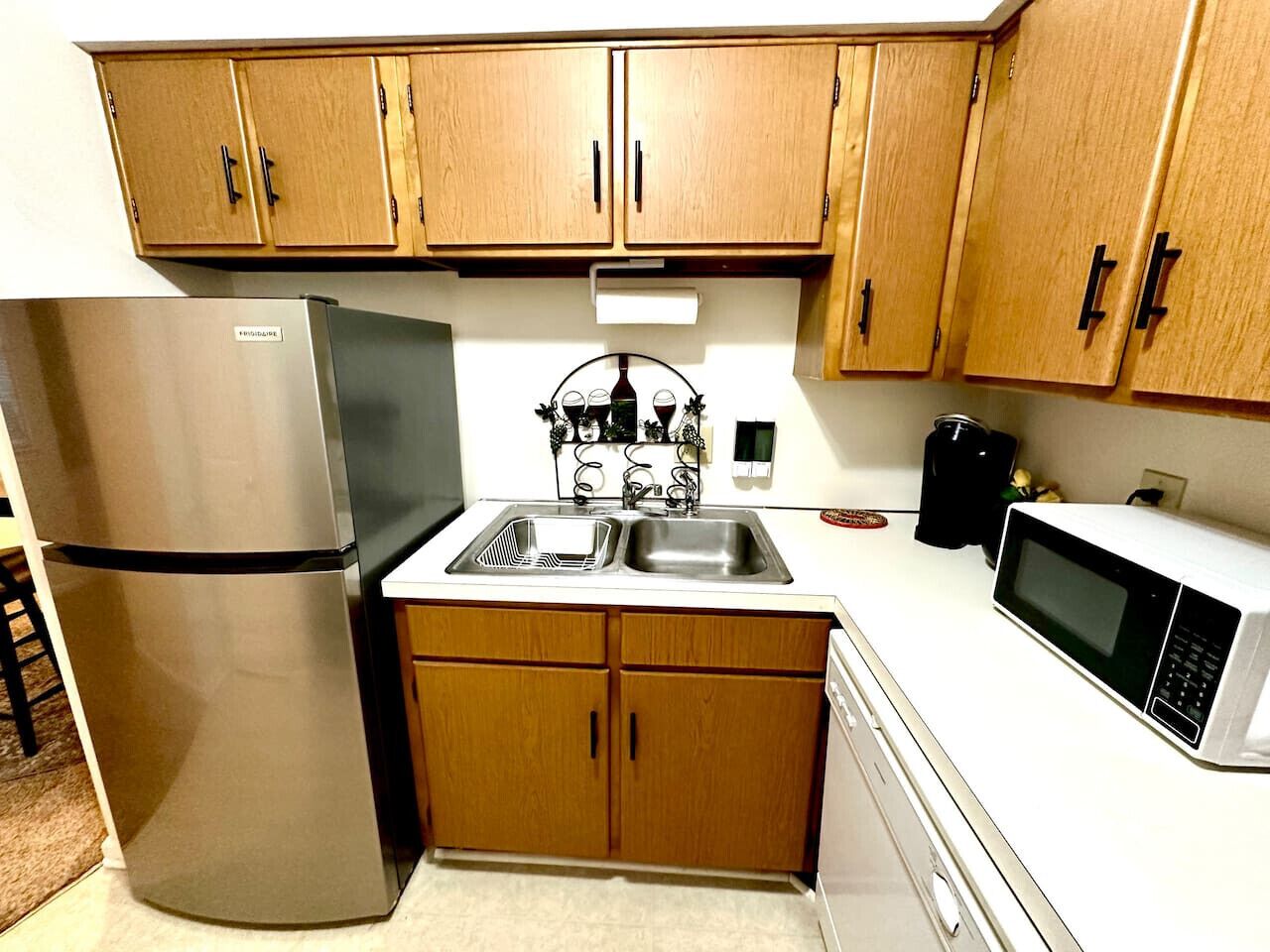 A compact kitchen with wooden cabinets, a stainless steel refrigerator, a double sink, and a microwave.