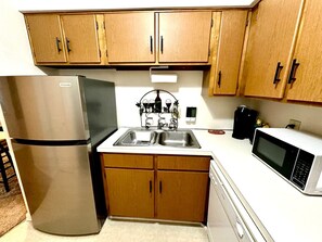 A compact kitchen with wooden cabinets, a stainless steel refrigerator, a double sink, and a microwave. - Cozy Downtown Destination (Raleigh)