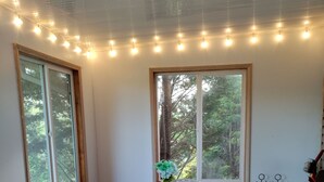 A dining area with a round table, white chairs, and a vase with flowers. There are two windows with curtains, a ceiling with string lights, and a wooden floor. - Private, cozy home - Handicapped Accessible no by- with great views  (Fletcher)