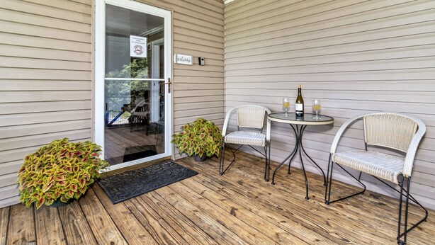 A covered porch with a sliding glass door, two wicker chairs, a small round table with a bottle and glass, and a welcome sign. - Private, cozy home - Handicapped Accessible no by- with great views  (Fletcher)