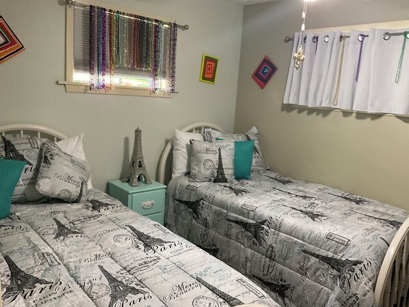 A bedroom with two beds, each with Paris-themed bedding. There is a small bedside table with a lamp, a window with decorative curtains, and wall art. - Charming water front cottage on Cane River with stunning sunroom & private decks (Natchitoches)
