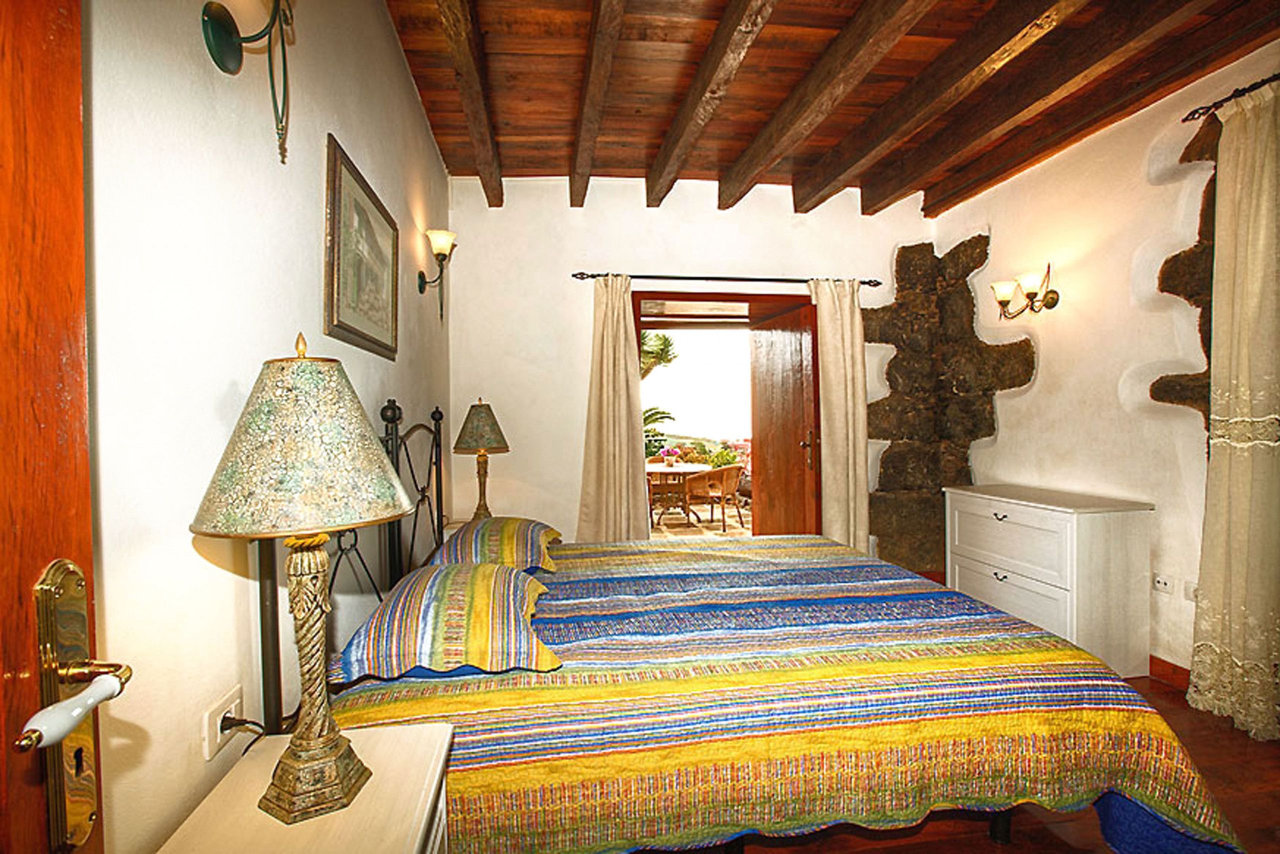 Une chambre à coucher avec un lit, une table de chevet, une lampe, une commode et un mur en pierre.