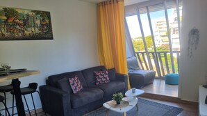 A modern living room with a black sofa, a dining table with chairs, a small coffee table, and a view of a balcony with a blue bean bag. - Very nice T2 40m2 + 45m2 terrace (La Grande-Motte)