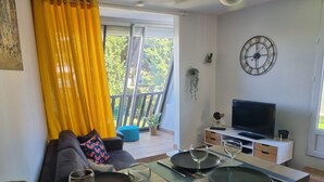 A modern living room with a sofa, a dining table set for two, a television, and a balcony with yellow curtains. - Very nice T2 40m2 + 45m2 terrace (La Grande-Motte)
