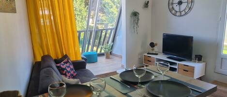 Un salon moderne avec un canapé, une table à manger pour deux personnes, un téléviseur et un balcon agrémenté de rideaux jaunes.