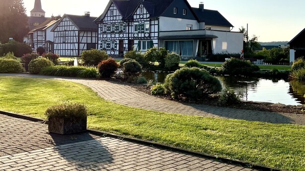 A traditional half-timbered house with a pond and a church spire in the background. - Vacation apartment Winterscheid - Barrier-free in the countryside - near Cologne/Bonn (Ruppichteroth)