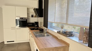 A modern kitchen with white cabinetry, a wooden countertop, stainless steel appliances, and a window with blinds. - Vacation apartment Winterscheid - Barrier-free in the countryside - near Cologne/Bonn (Ruppichteroth)