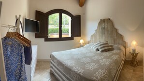 Ein Schlafzimmer mit einem Bett, zwei Nachttischlampen, einem an der Wand befestigten Fernseher, einem Fenster mit geöffneten Fensterläden und einem Kleiderschrank mit darin hängender Kleidung.