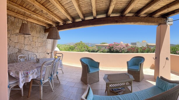 Eine überdachte Terrasse mit Steinwand, Tisch mit Stühlen und Korbmöbeln.
