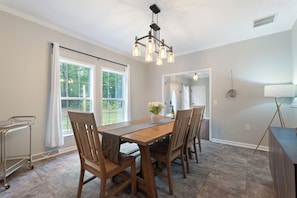 A dining area with a wooden table and chairs, a chandelier, and a view of the outdoors through large windows. - 6BR • Arcade & Pool Table • Hot Tub • Wine Country (Faber)