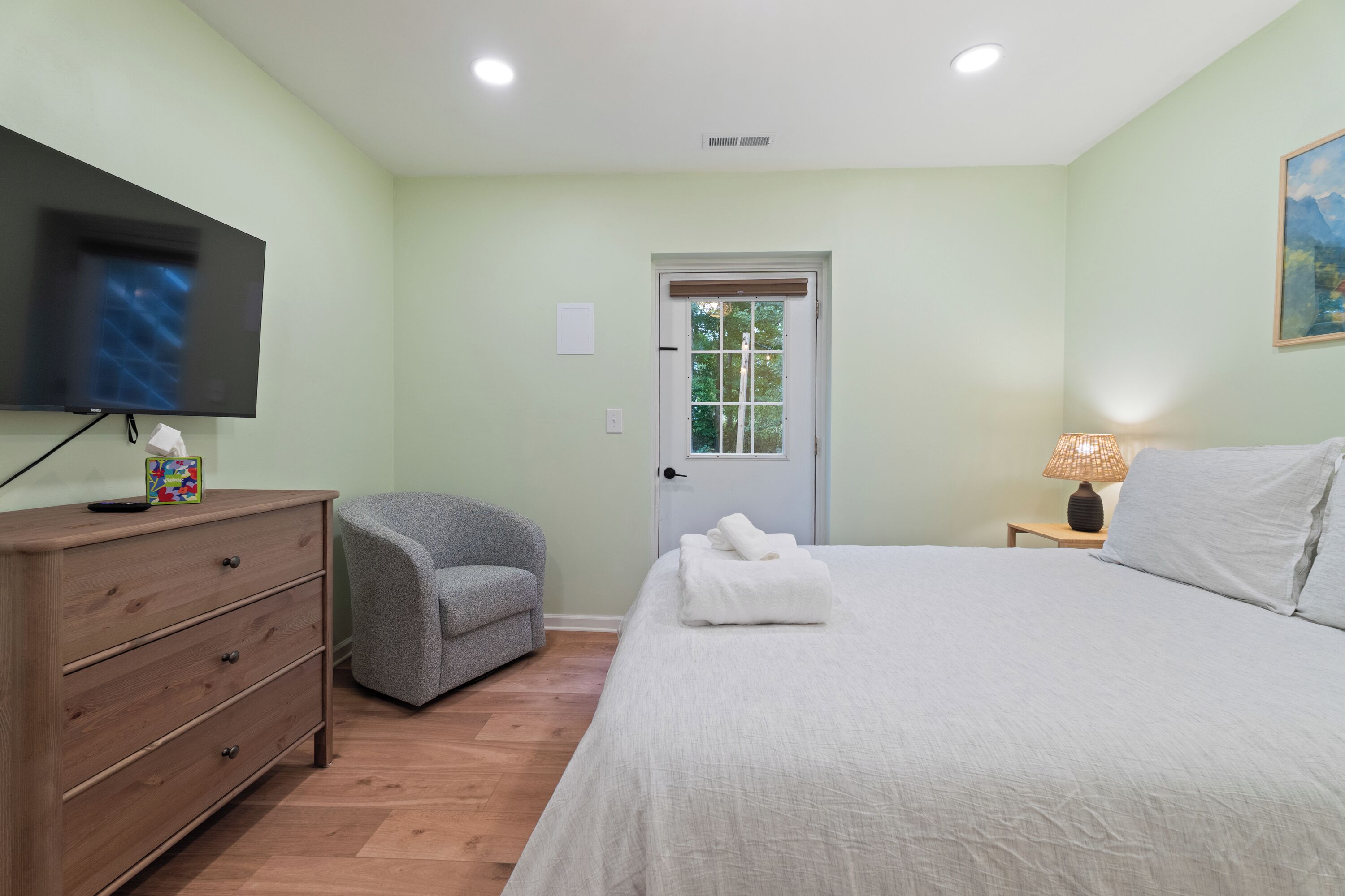 Un dormitorio con cama, cómoda, silla, televisor y puerta con ventanal.