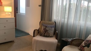 Una habitación acogedora con un sillón blanco, una alfombra redonda también blanca, una silla de madera con un cojín estampado, una mesita con una lámpara y vistas a través de una ventana con cortinas traslúcidas.