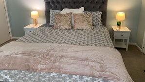 Un dormitorio con cabecera capitonada, ropa de cama estampada, dos mesitas de noche con lámparas y suelo alfombrado.