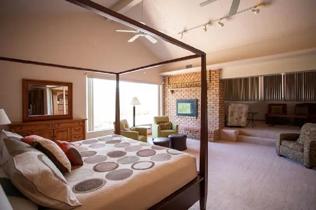 A four-poster bed with a patterned bedspread, a wooden dresser, a green armchair, and a brick wall with a television.