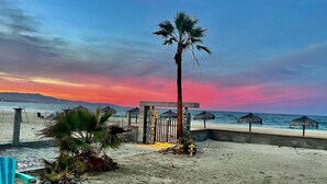 Escena de playa con una palmera, un portón de madera y sombrillas.