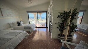 Habitación de hotel con dos camas, una planta grande en maceta y vista a la playa a través de la puerta corrediza de vidrio.