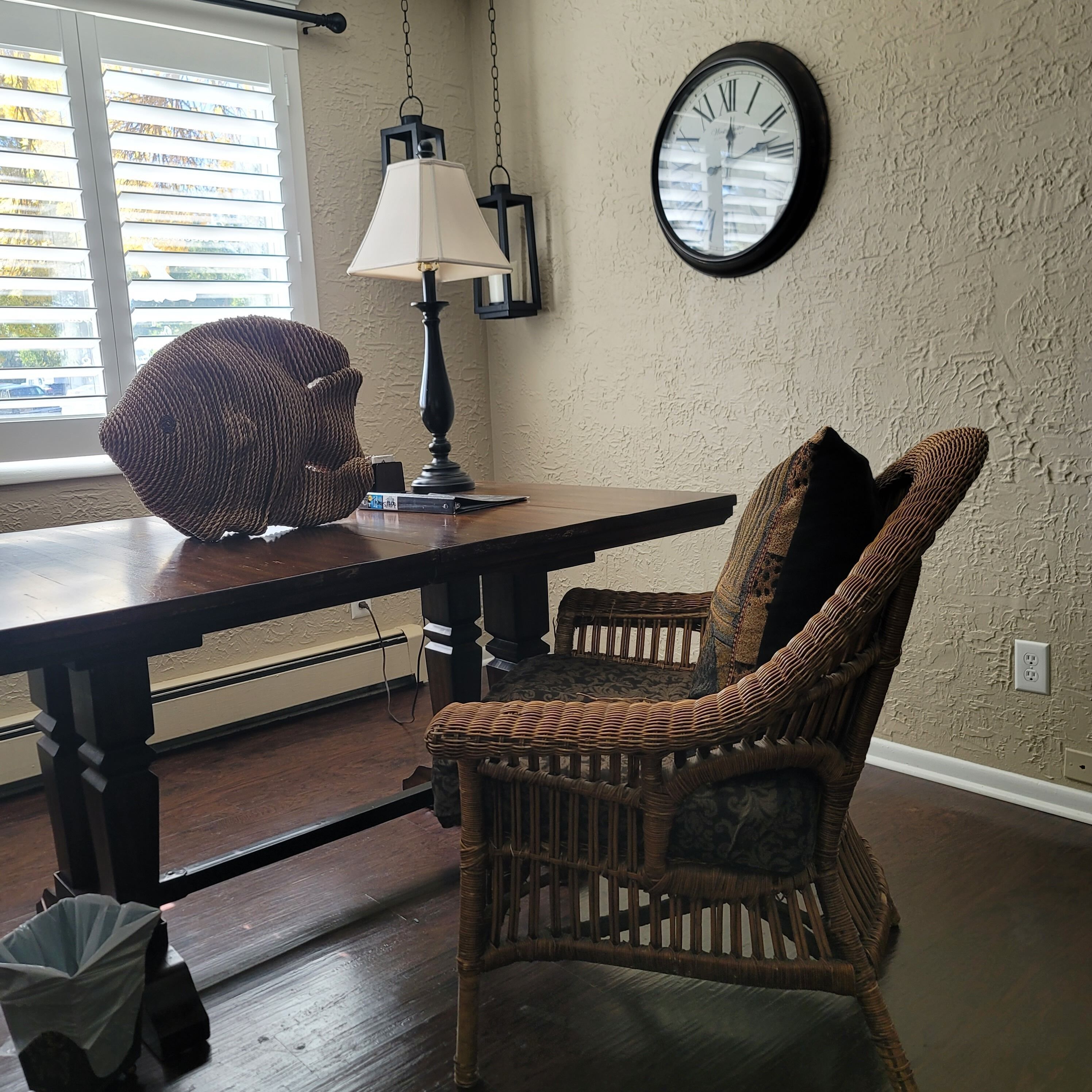 A dining area with a wooden table, wicker chairs, a wall clock, and a hanging lamp.