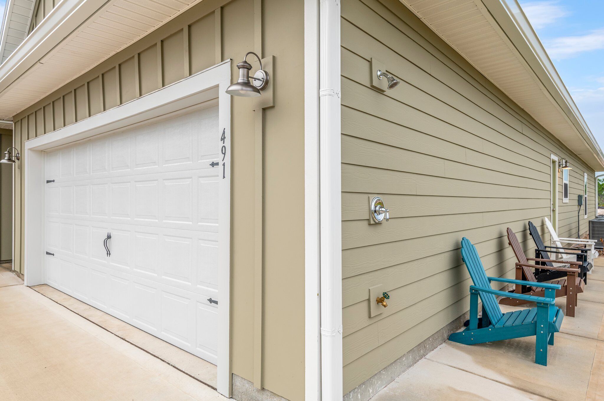 A beige house with a white garage door, outdoor lighting, and Adirondack chairs.
