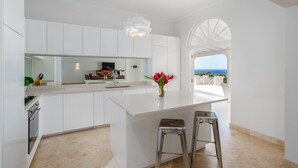 Une cuisine moderne avec des meubles blancs, un grand îlot central doté de tabourets, un luminaire suspendu et une vue sur la mer, visible à travers une porte en arc de cercle.