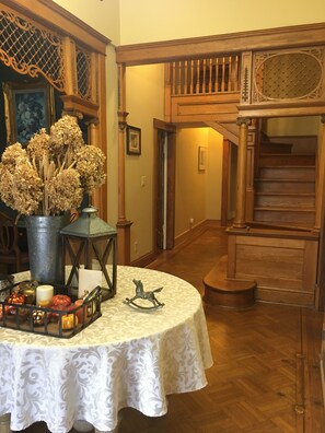 A room with a round table covered in a white tablecloth, a vase with dried flowers, a lantern, and a small rocking horse ornament. A staircase with wooden railings and a wooden door is visible in the background.
