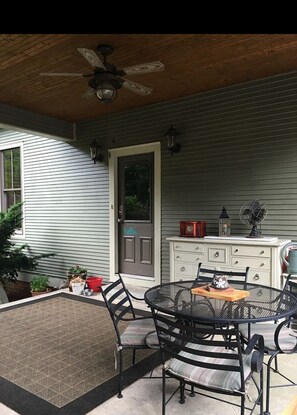 Une terrasse couverte avec une table et des chaises rondes en métal, un ventilateur de plafond et une commode blanche agrémentée d’objets décoratifs.