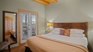 Un dormitorio con cabecera de madera, cama con ropa de cama blanca, una banca, una puerta con vistas y una lámpara en una mesita de noche.